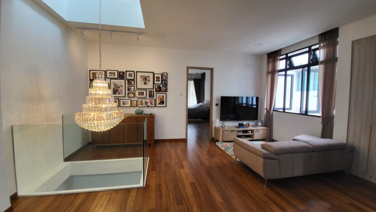 A modern living room with a large chandelier, photo gallery wall, corner sofa, TV on a stand, and wooden flooring. A staircase with glass railing is partially visible.